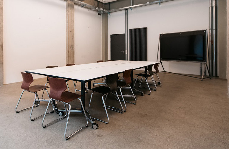 Foto: Moderner Seminarraum mit weißen Tischen, braunen Stühlen und einem großen Bildschirm.