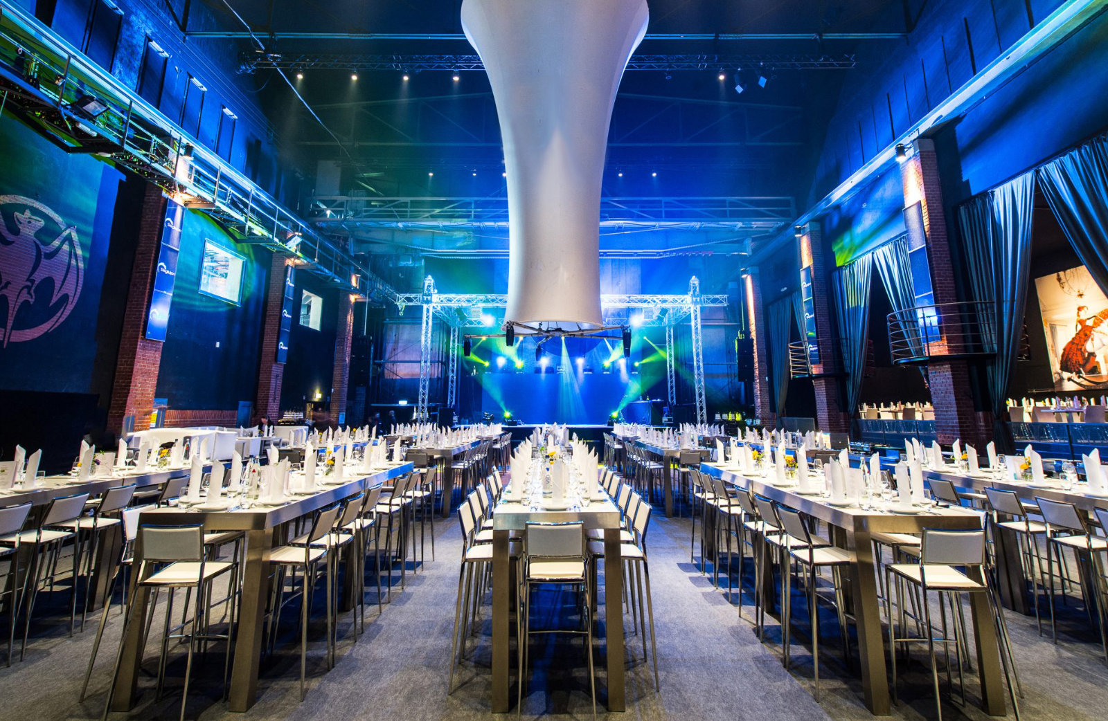 Foto: Großer Veranstaltungsraum mit hohen Decken, langen gedeckten Tischen, blauer Beleuchtung und Bühne.